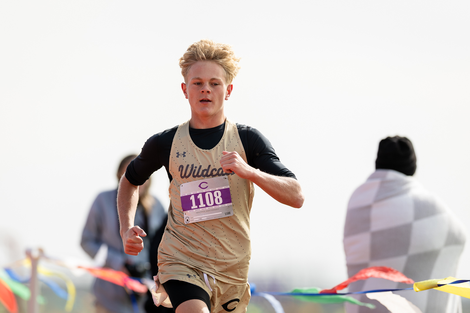 3A-4A-district-xc-jpegJoe-pps-97 | Press Pass Sports Young male runner in a beige Wildcats singlet with bib 1108, sprinting on a course lined with colorful banners.