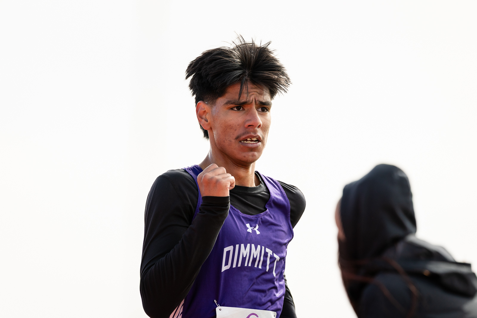 3A-4A-district-xc-jpegJoe-pps-99 | Press Pass Sports Male runner in a purple DIMITT singlet raises a clenched fist, mid-race, with a blurred hoodie-clad competitor nearby.