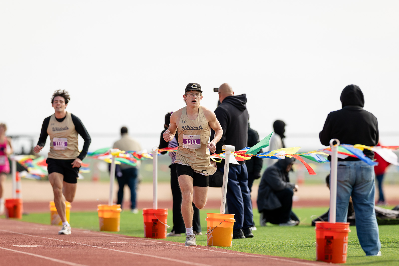 3A-4A-district-xc-jpegJoe-pps-101 | Press Pass Sports Male athlete in a beige Wildcats singlet and cap sprinting on a track, with spectators and colorful banners in the background.