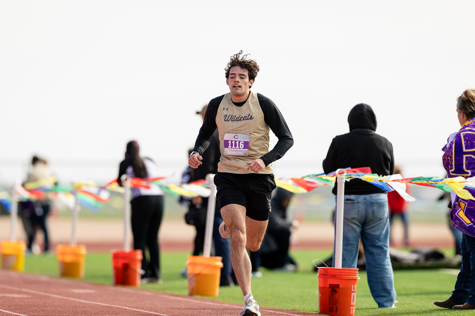 3A-4A-district-xc-jpegJoe-pps-102 | Press Pass Sports Male runner in beige Wildcats jersey crossing the track finish line, bib 1116, with cheering spectators and colorful finish-line ribbons in the background.