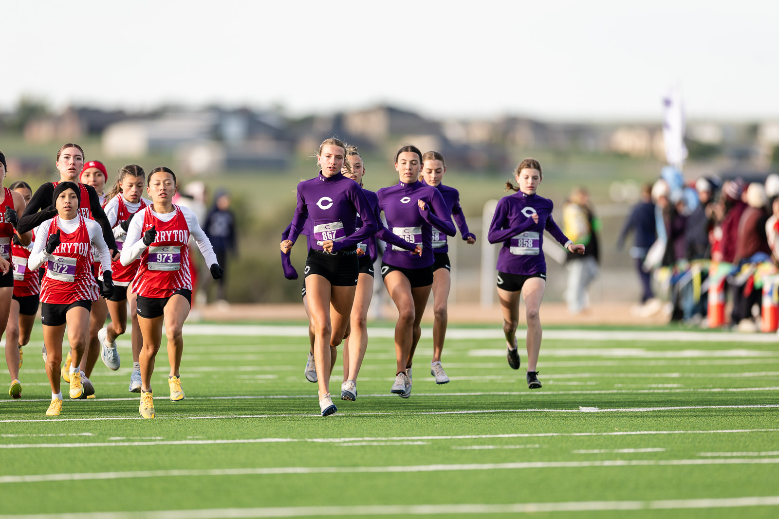 canyon-eagles-district-xc-jpegJoe-pps-1 | Press Pass Sports Girls' cross-country team race on a green track, runners in red and purple uniforms sprint toward the finish with spectators in the background.