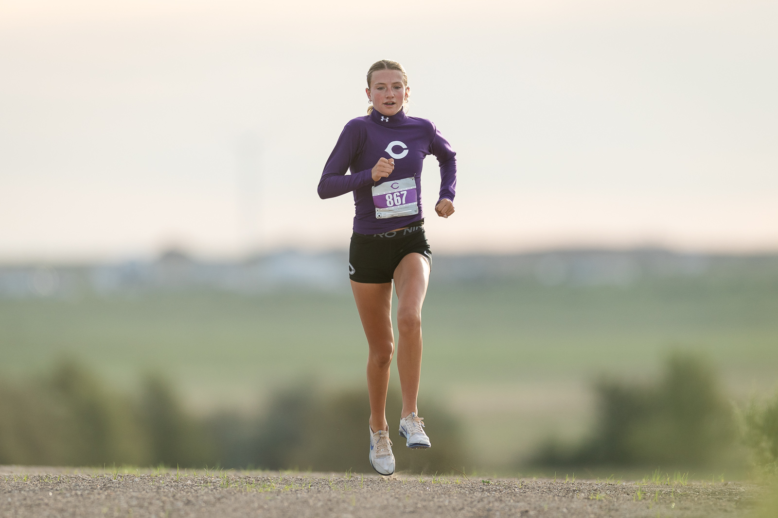 canyon-eagles-district-xc-jpegJoe-pps-2 | Press Pass Sports Female runner in a purple long-sleeve top and black shorts leaps mid-air during a race on an open countryside road.