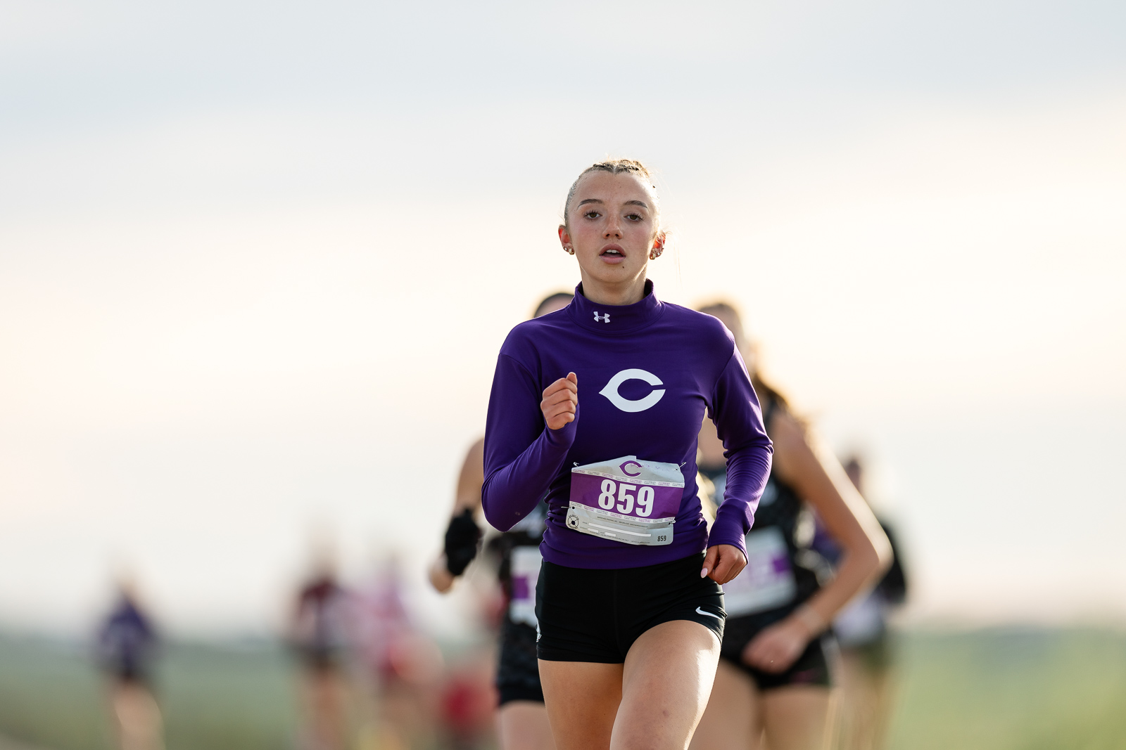 canyon-eagles-district-xc-jpegJoe-pps-3 | Press Pass Sports Female runner in a purple long-sleeve top with bib 859, running in a race at sunset with other runners blurred behind her.