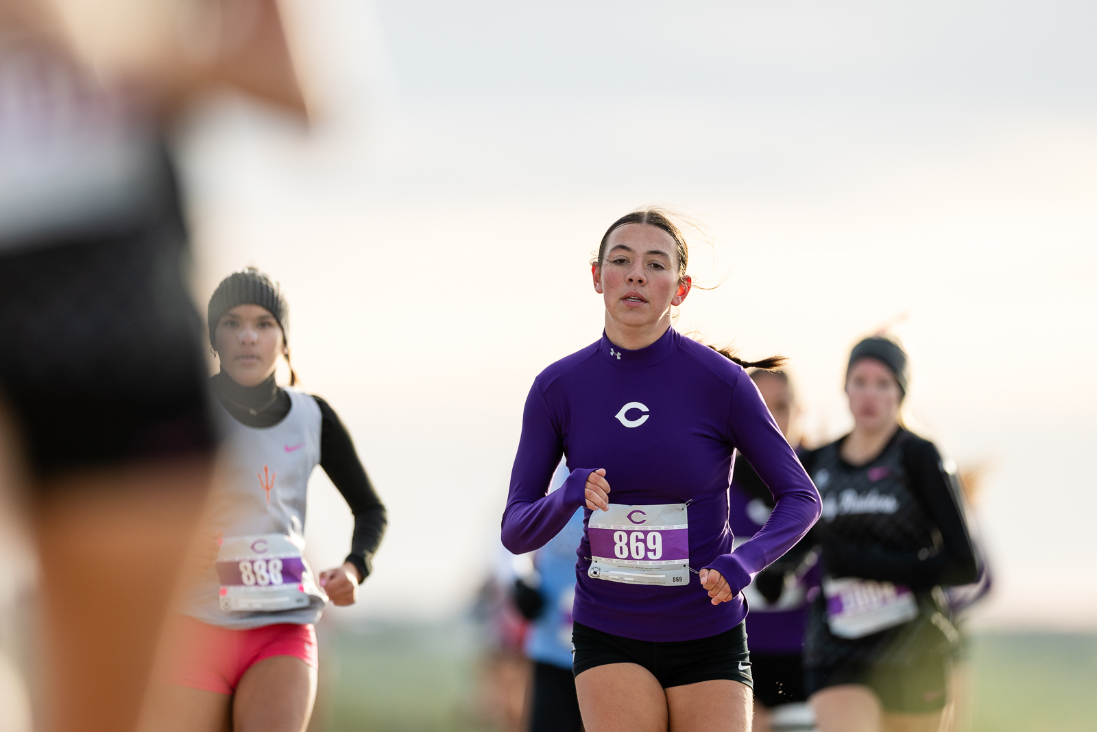 canyon-eagles-district-xc-jpegJoe-pps-4 | Press Pass Sports Female runner in a purple long-sleeve top with bib 869 leading a race, others behind on a sunny day
