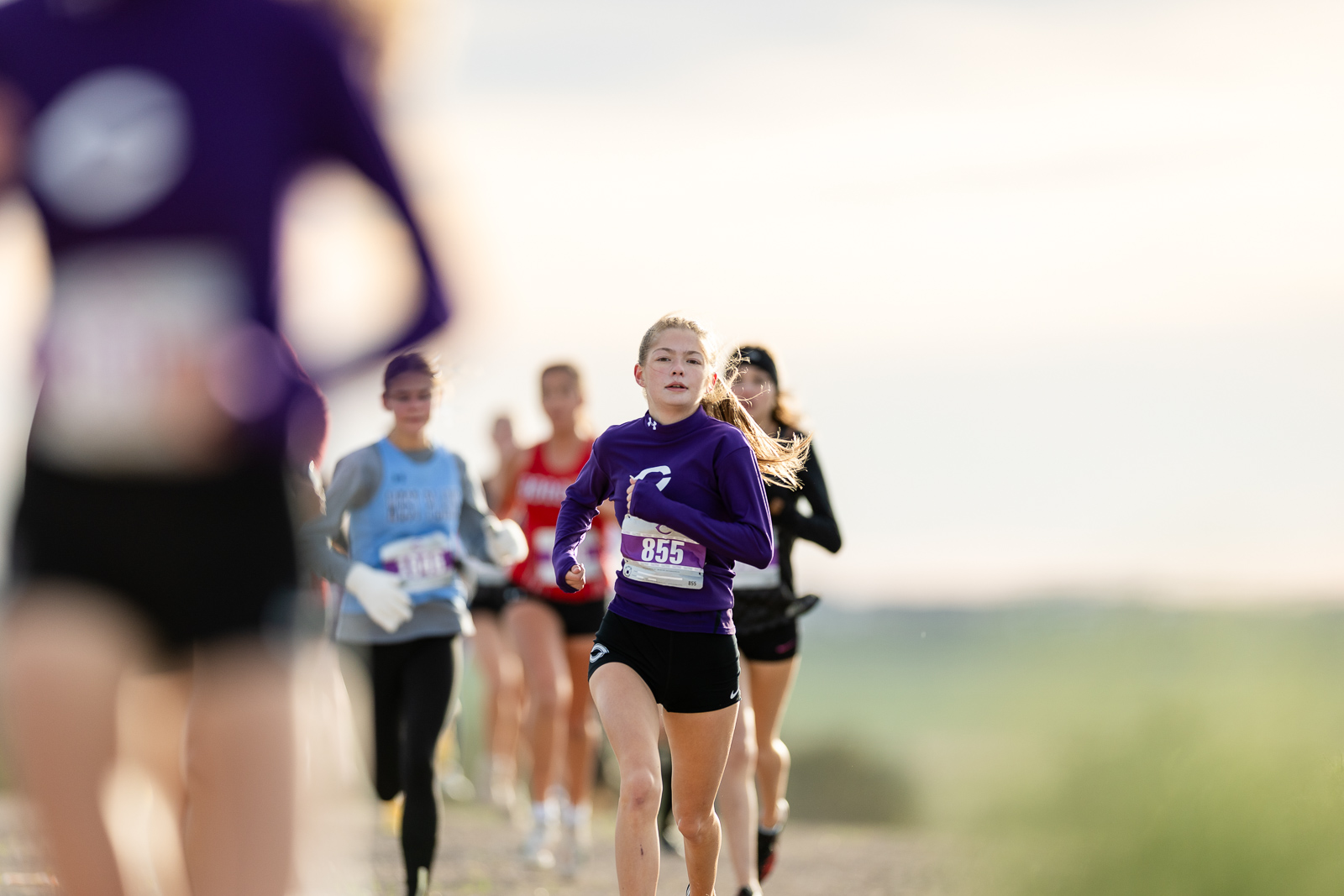 canyon-eagles-district-xc-jpegJoe-pps-6 | Press Pass Sports Young girl in a purple long-sleeve top runs in a road race, bib 855, with other runners blurred in the background on a sunny day.