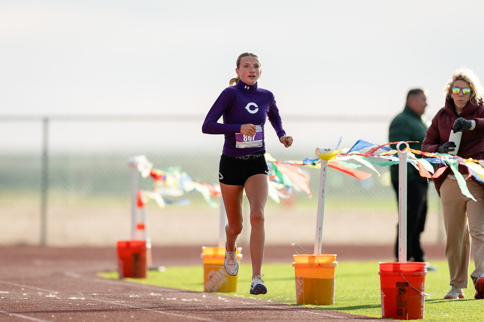 canyon-eagles-district-xc-jpegJoe-pps-7 | Press Pass Sports Female sprinter in a purple long-sleeve top crosses a track finish line, with colorful ribbons and spectators in the background.
