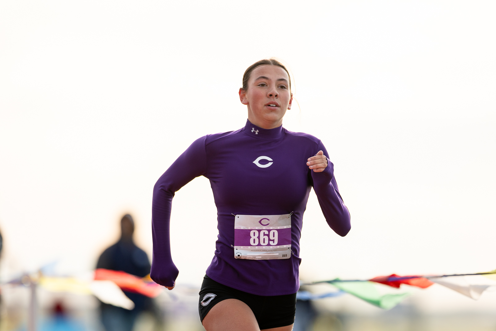 canyon-eagles-district-xc-jpegJoe-pps-8 | Press Pass Sports Female runner in a purple long-sleeve top with bib 869 sprinting during a race, outdoors with a bright background.