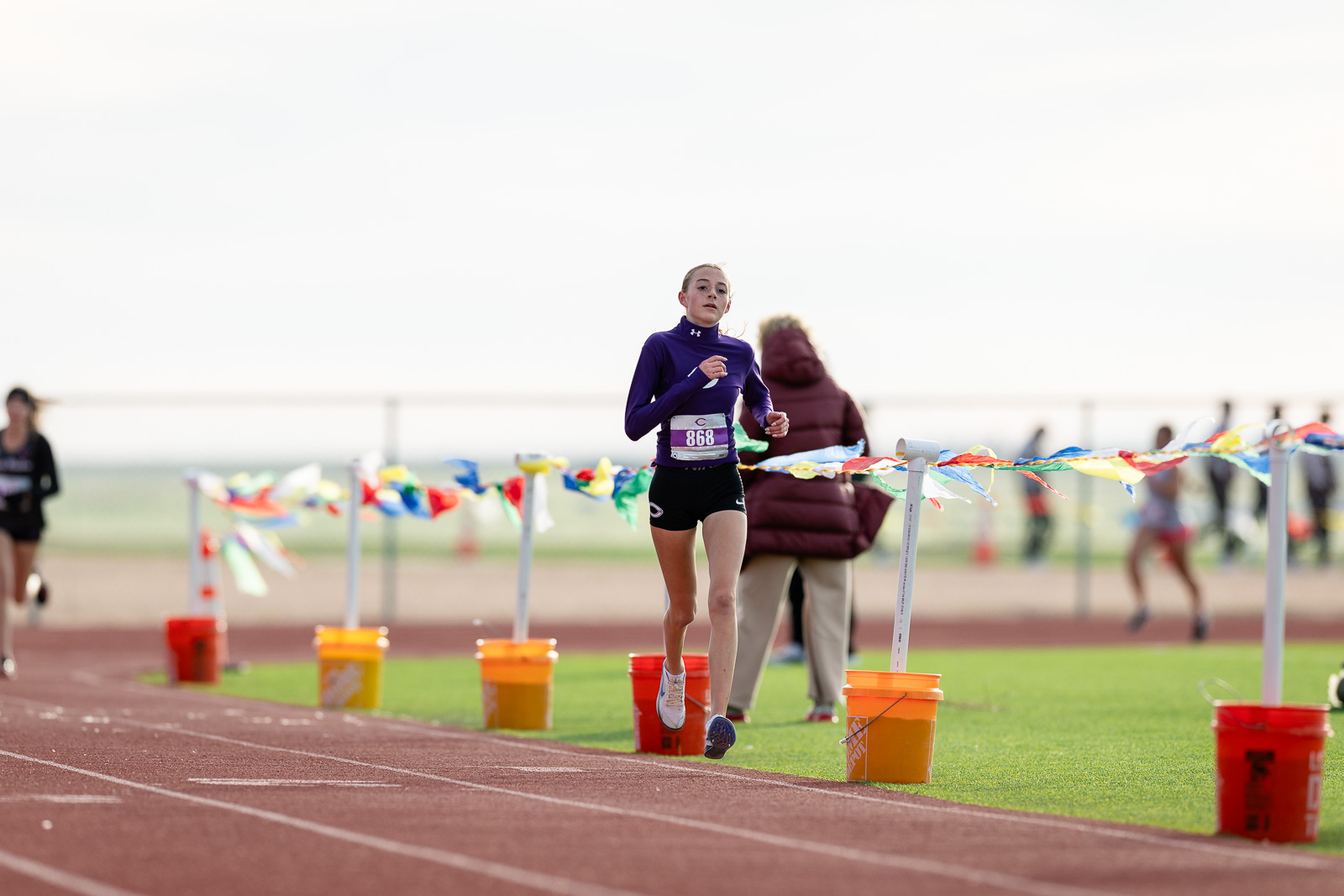 canyon-eagles-district-xc-jpegJoe-pps-9 | Press Pass Sports Female sprinter in a purple long-sleeve top and black shorts runs on a track, bib 868, with colorful finish-line flags in the background.