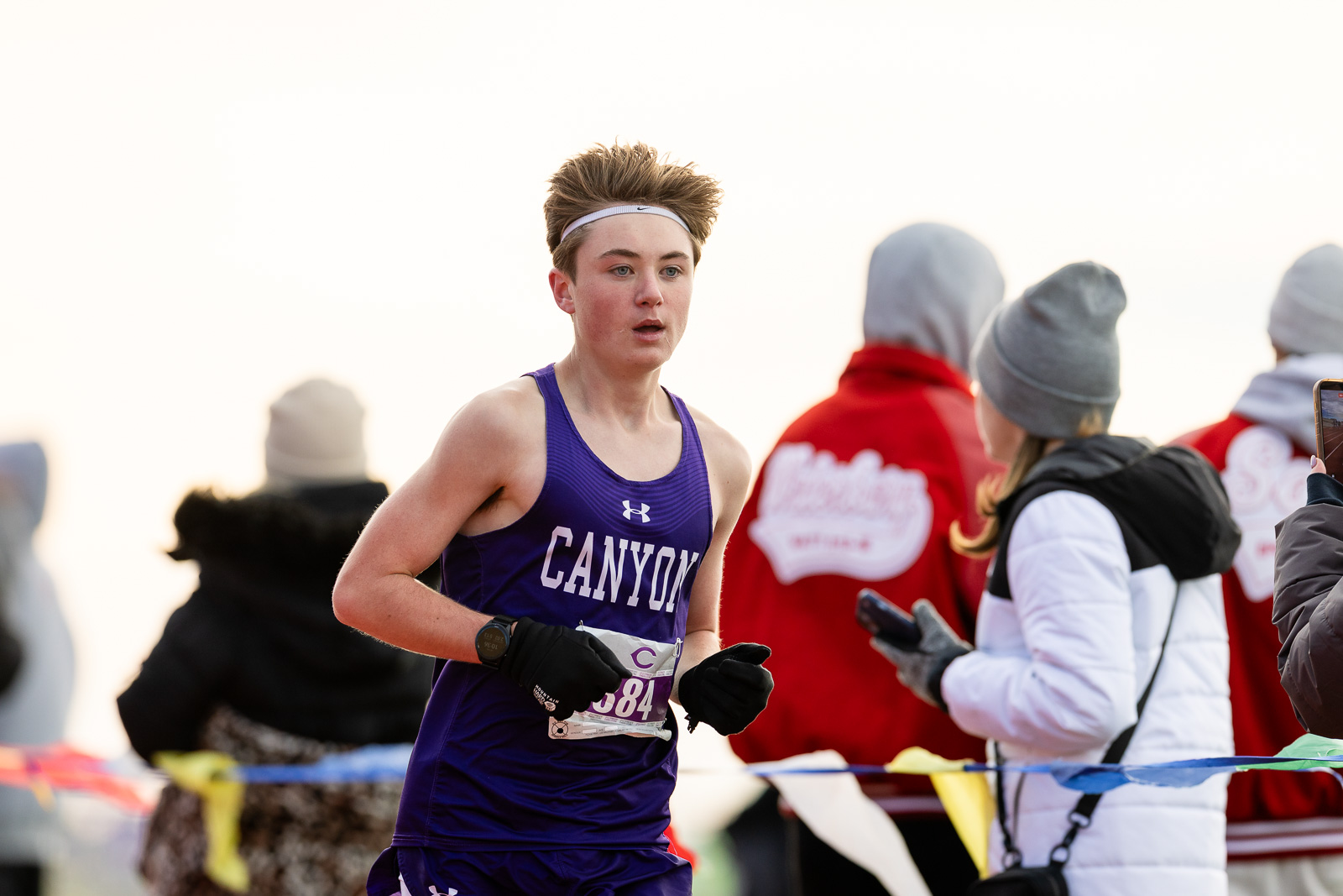 canyon-eagles-district-xc-jpegJoe-pps-14 | Press Pass Sports Female cross-country runner in a purple Canyon uniform with a white headband, mid-race with onlookers and finish-line tape in the background.