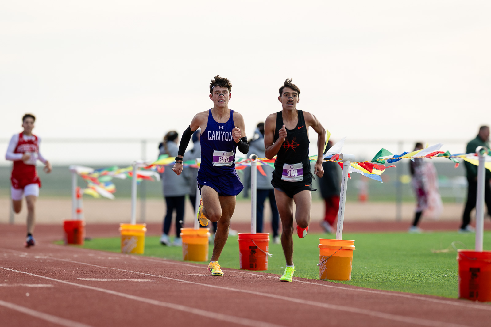 canyon-eagles-district-xc-jpegJoe-pps-16 | Press Pass Sports Two male sprinters lead a track race toward the finish, one in a blue Canyon uniform (886) and the other in black (900), with colorfully marked lanes behind them.