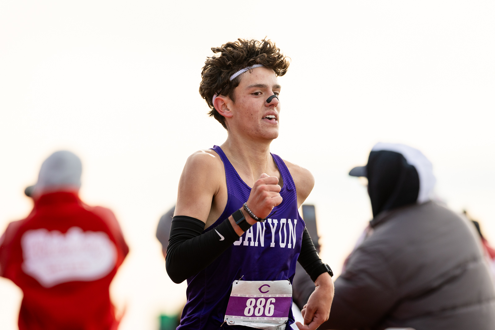 canyon-eagles-district-xc-jpegJoe-pps-17 | Press Pass Sports Male runner in a purple singlet with bib 886, mid-stride in a race, flanked by cheering spectators in jackets.