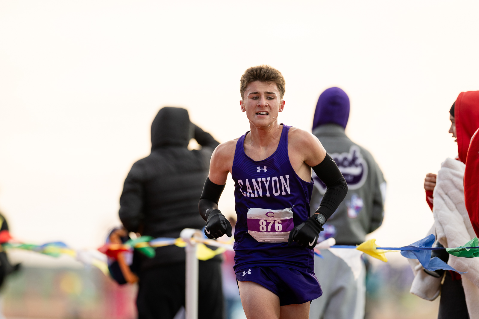 canyon-eagles-district-xc-jpegJoe-pps-18 | Press Pass Sports Young male runner in a purple Canyon singlet and shorts, racing with bib 876, gritting his teeth as he runs along a course lined with spectators.
