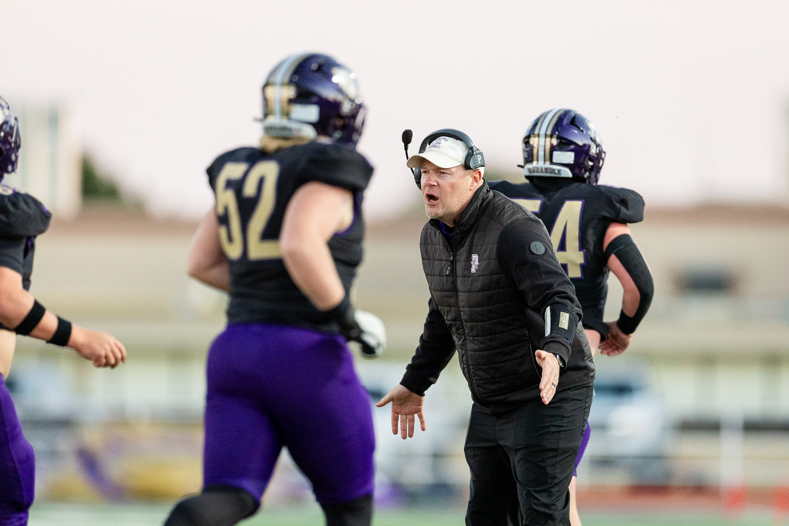 spearman-vs-panhandle-football-jpegJoe-pps-6 | Press Pass Sports Football coach in a black vest shouts instructions to players during a football practice on the field, wearing a headset and cap.
