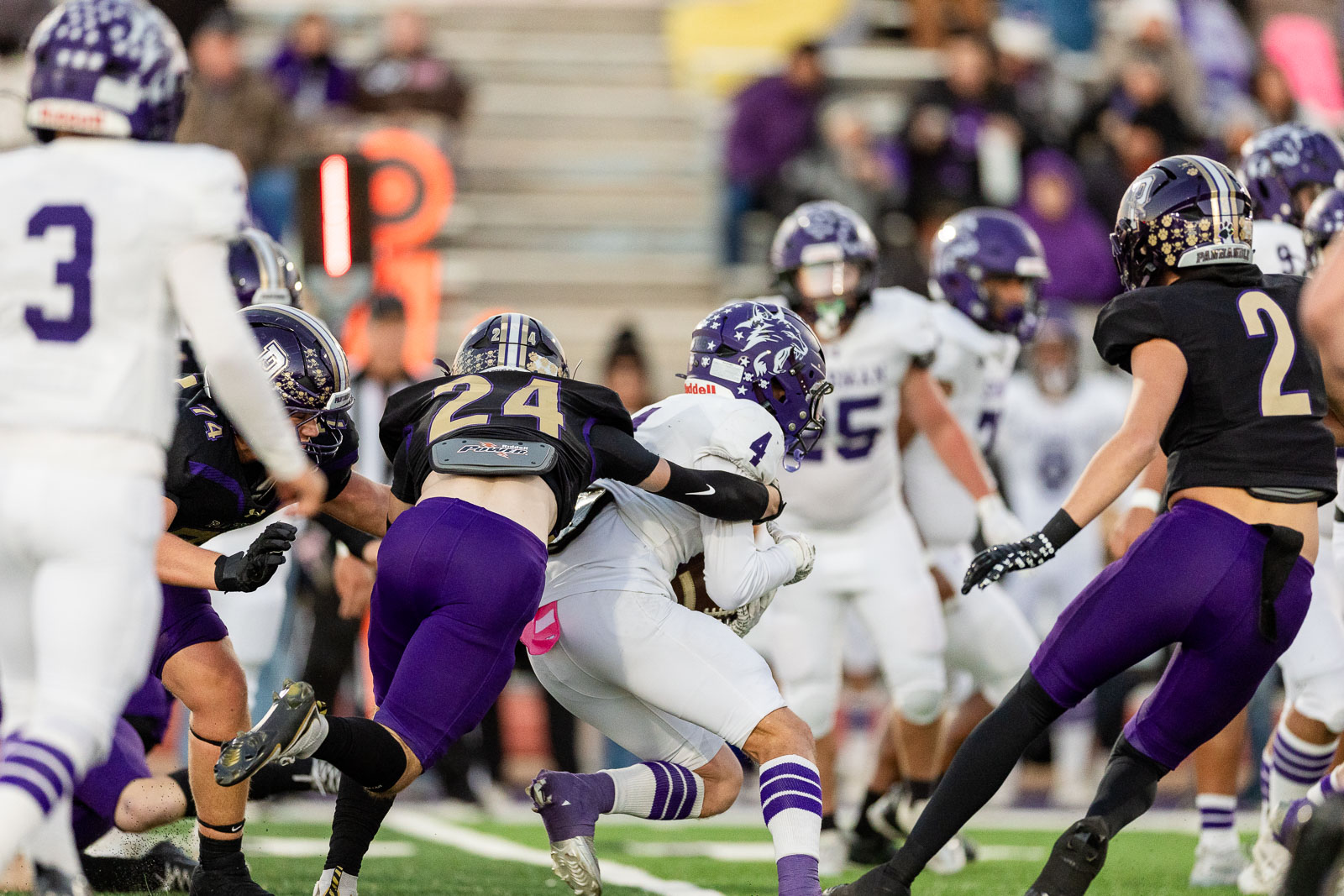 spearman-vs-panhandle-football-jpegJoe-pps-7 | Press Pass Sports Players collide during a football game; a white-uniformed carrier is tackled by a purple-uniformed defender as the crowd watches from the stands in the background (green turf).