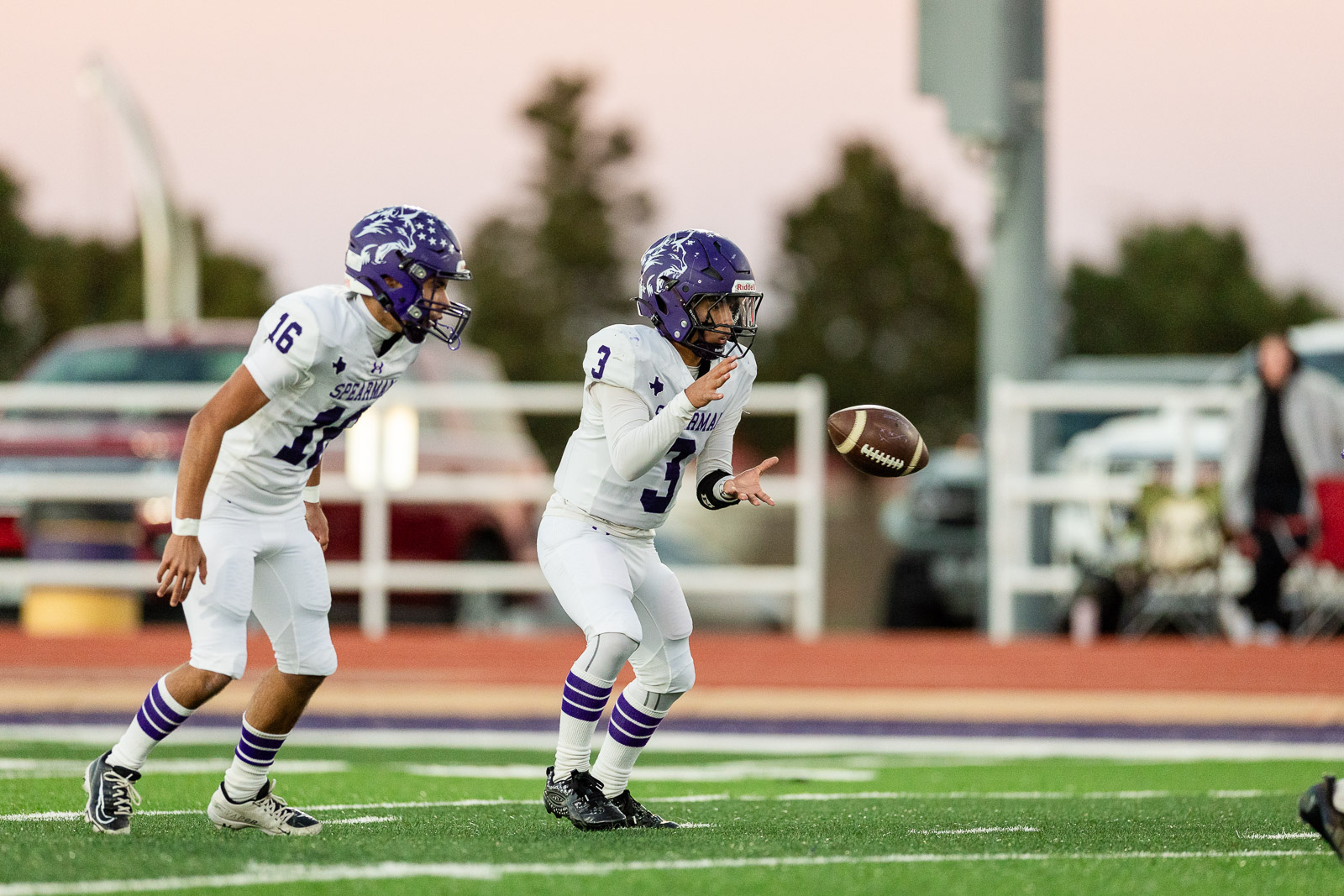 spearman-vs-panhandle-football-jpegJoe-pps-8 | Press Pass Sports Two football players in white and purple uniforms chase the ball on a green turf during a game; one player (#3) reaches for the ball as another (#16) closes in.