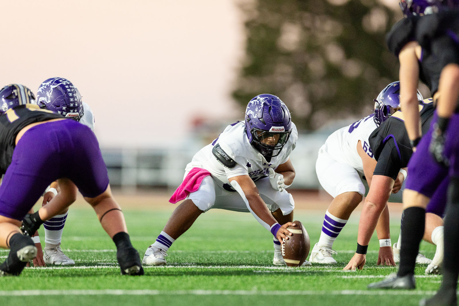 spearman-vs-panhandle-football-jpegJoe-pps-10 | Press Pass Sports Football players in white and purple line up at the line of scrimmage as the center snaps the ball, one player crouched low to receive it on a sunny field.