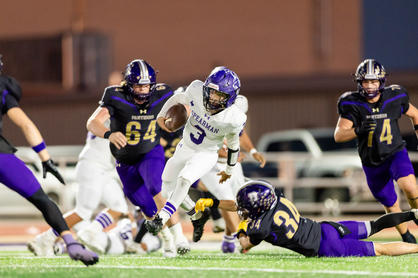 spearman-vs-panhandle-football-jpegJoe-pps-15 | Press Pass Sports Football player in white Spearman jersey runs with the ball while defenders in purple/black chase him on a green field stadium lights