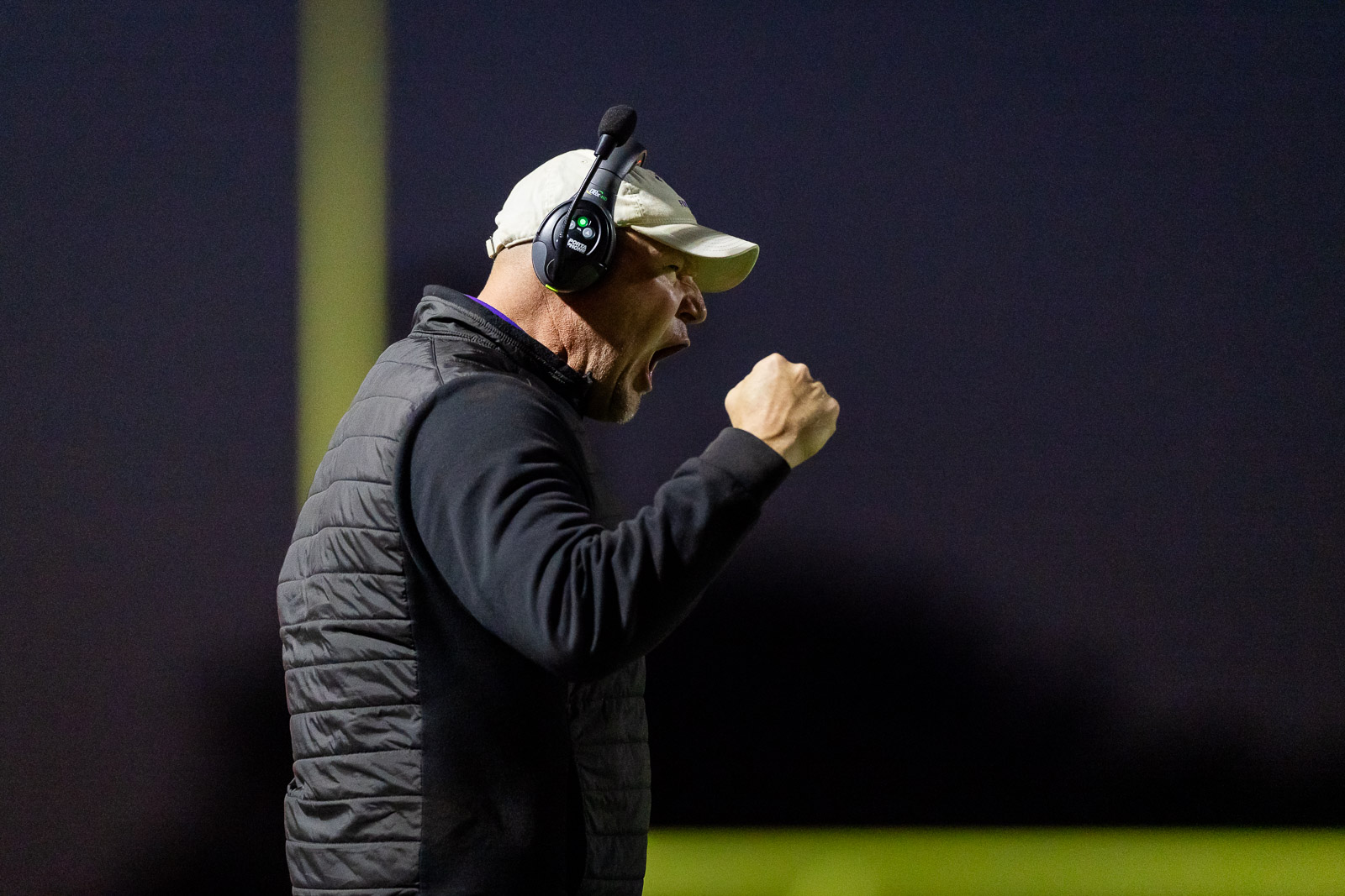 spearman-vs-panhandle-football-jpegJoe-pps-17 | Press Pass Sports Coach wearing a beige cap and headset, fist raised in celebration on a dimly lit football field at night.