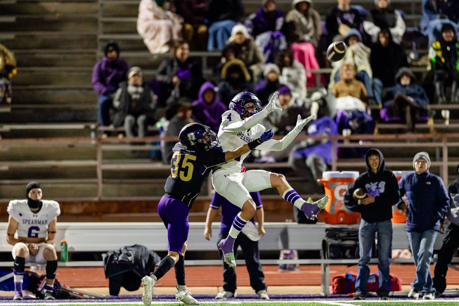spearman-vs-panhandle-football-jpegJoe-pps-18 | Press Pass Sports Two football players leap for the ball as a pass sails overhead, while spectators watch from the bleachers in a night game.