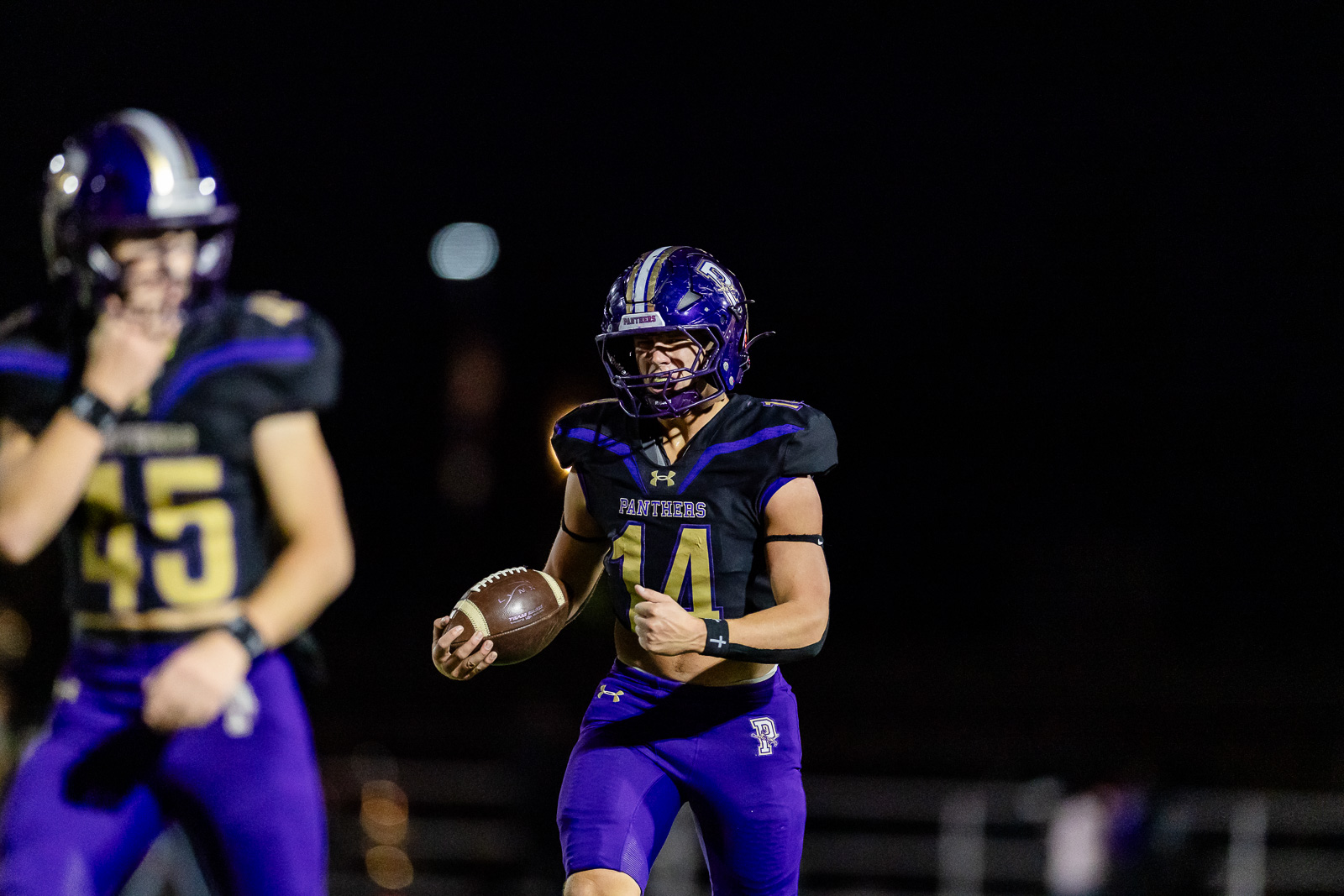 spearman-vs-panhandle-football-jpegJoe-pps-19 | Press Pass Sports Night-time football game: a Panther player in #14 black and purple uniform runs with the ball, focused and mid-stride, stadium lights in the background