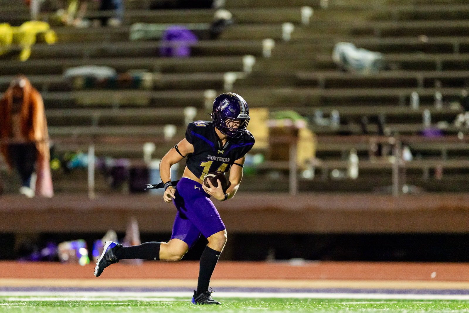 spearman-vs-panhandle-football-jpegJoe-pps-20 | Press Pass Sports A football player in a purple and black uniform runs with the ball on a field, looking ahead during a night game.