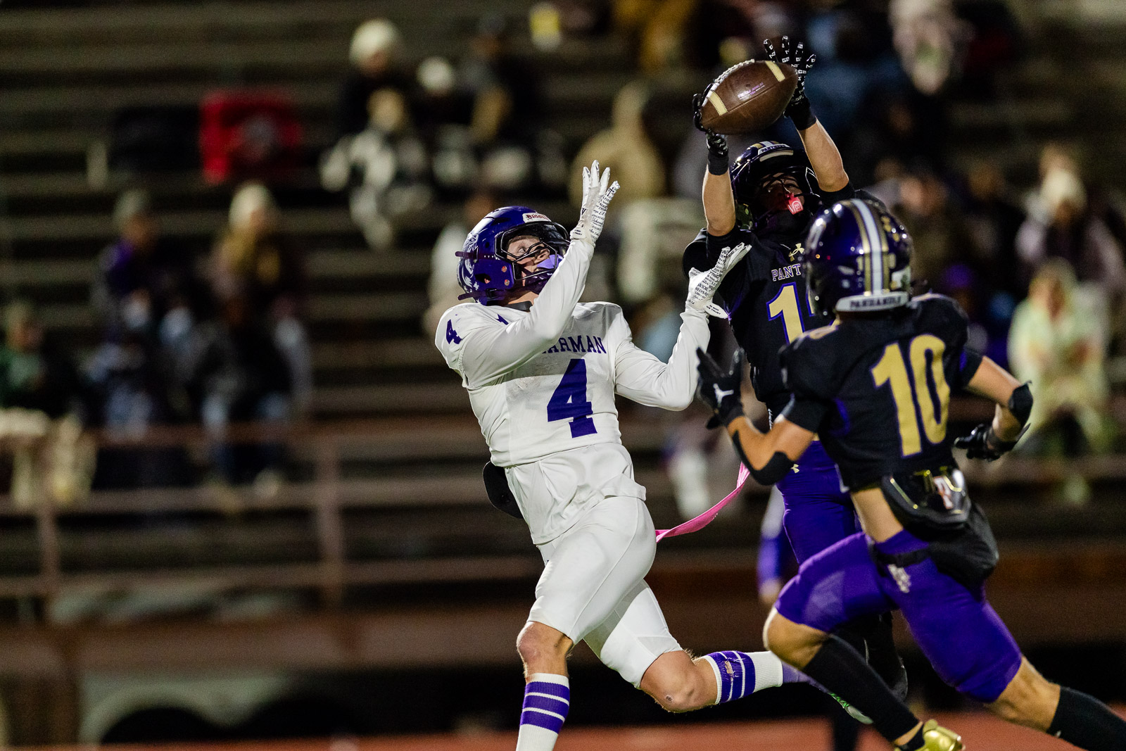 spearman-vs-panhandle-football-jpegJoe-pps-21 | Press Pass Sports Football players leap for a catch; white team #4 reaches up as a dark-uniform opponent defends, ball in the air under stadium lights.