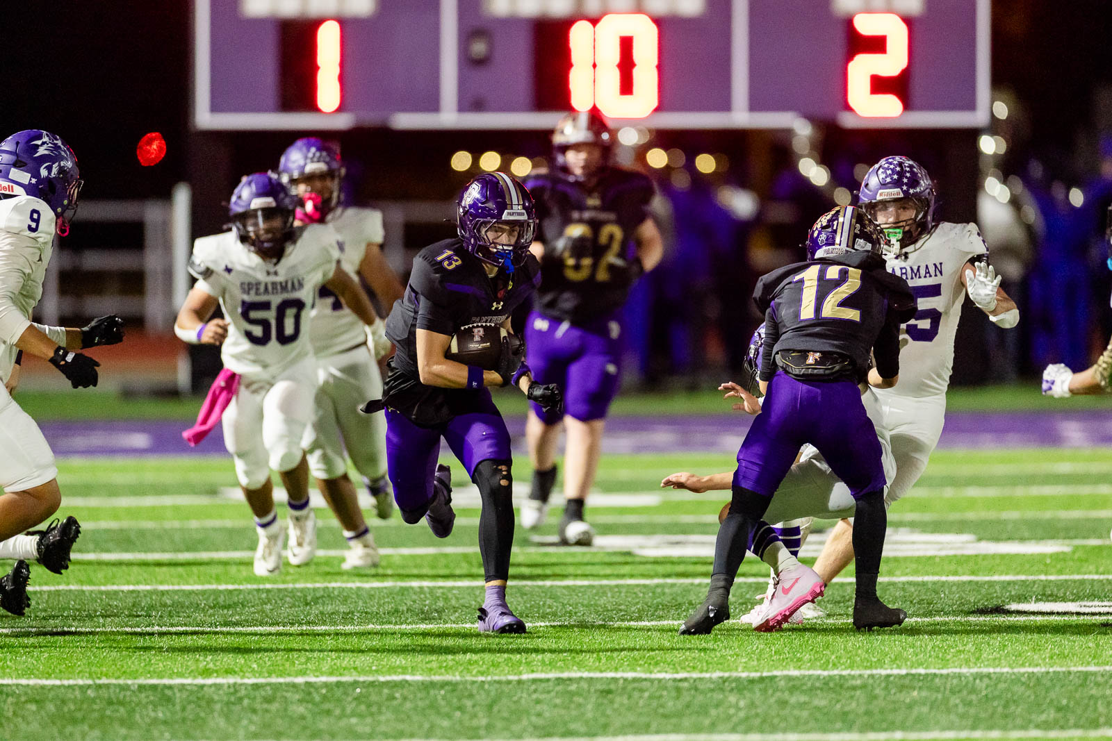 spearman-vs-panhandle-football-jpegJoe-pps-22 | Press Pass Sports A football player in a dark purple uniform runs with the ball while white-uniformed players chase on a bright, artificial turf field at night, with a large scoreboard behind them showing the score.”,