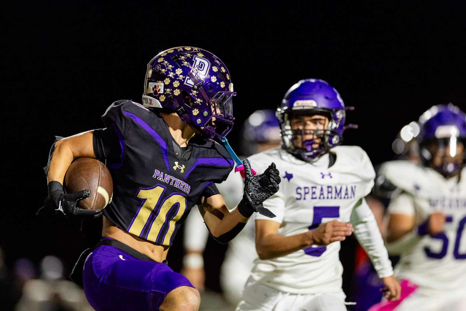 spearman-vs-panhandle-football-jpegJoe-pps-23 | Press Pass Sports Nighttime football action: a Panthers player in purple and gold (number 10) runs with the ball as defenders in white Spearman uniforms close in, under stadium lights.