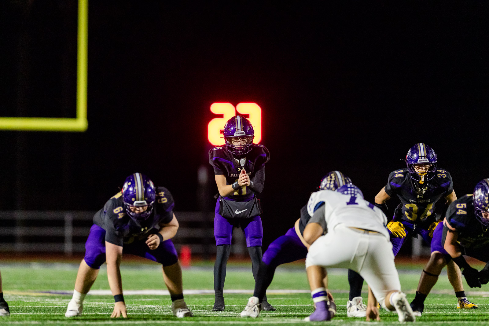 spearman-vs-panhandle-football-jpegJoe-pps-24 | Press Pass Sports High school football players line up at the snap on a night game; quarterback with hands together center stage, teammates in purple and black uniforms.