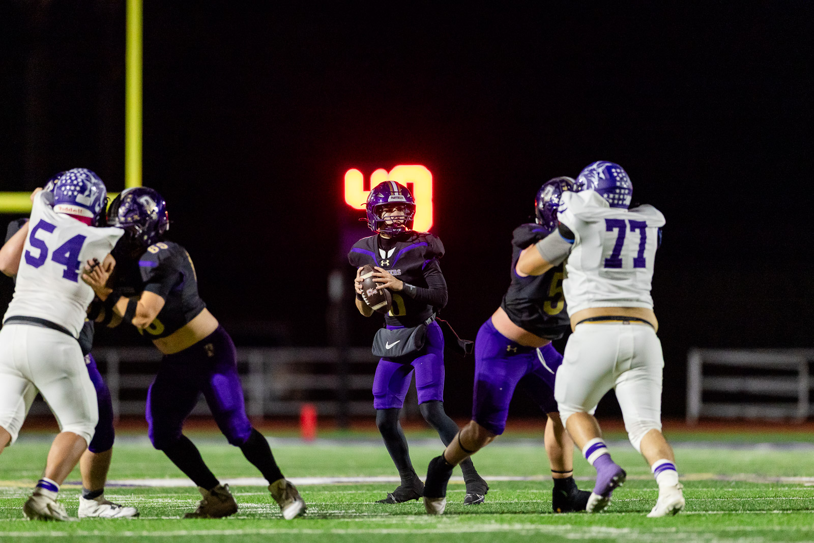 spearman-vs-panhandle-football-jpegJoe-pps-25 | Press Pass Sports Quarterback in purple with ball, scanning field as teammates block during a nighttime football game.