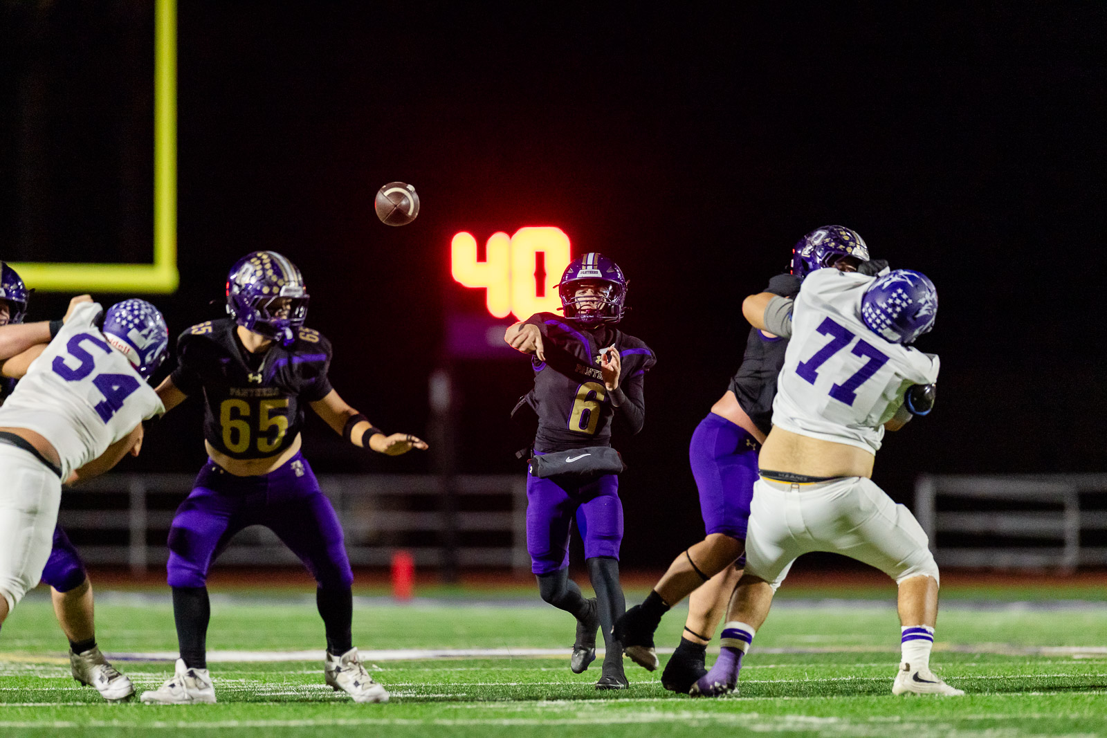 spearman-vs-panhandle-football-jpegJoe-pps-26 | Press Pass Sports Quarterback in purple (#6) throws a pass during a night game as linemen and defenders converge nearby on the field.