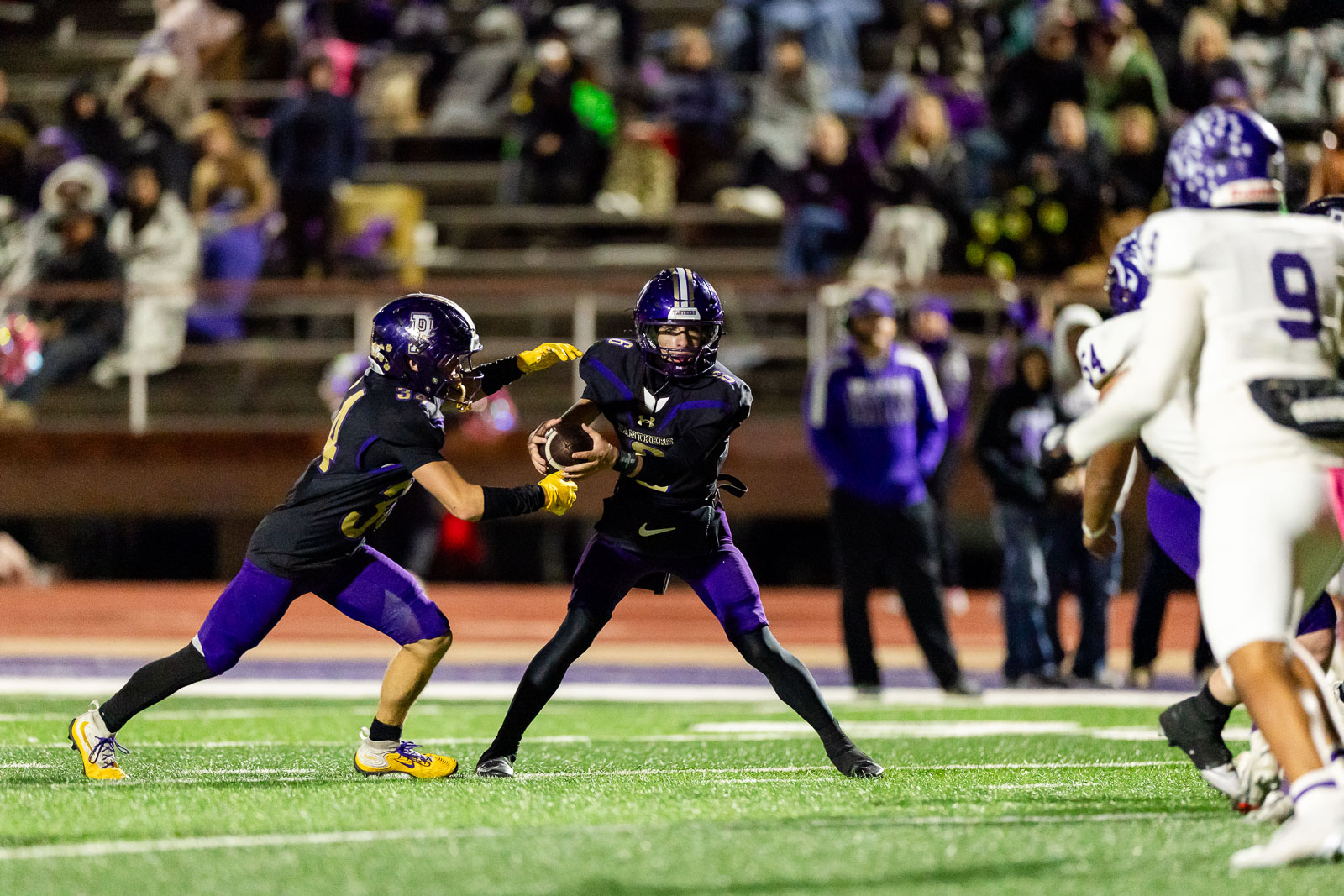 spearman-vs-panhandle-football-jpegJoe-pps-30 | Press Pass Sports A football player in purple and black holds the ball as a teammate blocks, on a turf field with spectators in the stands behind them.