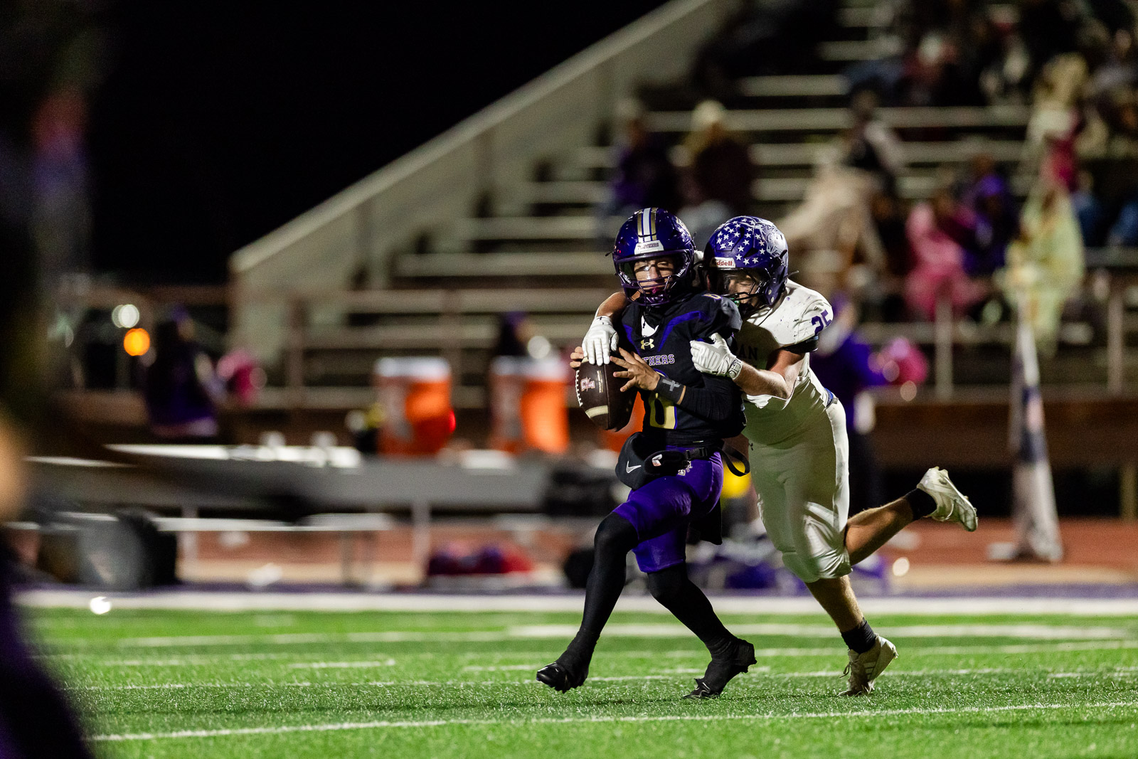 spearman-vs-panhandle-football-jpegJoe-pps-31 | Press Pass Sports Football player in a purple and black uniform runs with the ball while a white-team defender reaches in during a night game on a lit turf field.