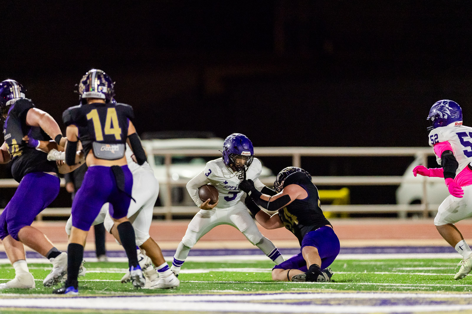 spearman-vs-panhandle-football-jpegJoe-pps-32 | Press Pass Sports Football player in a white jersey and purple helmet carries the ball while a defender in black purple dives in during a night game on a bright green field.