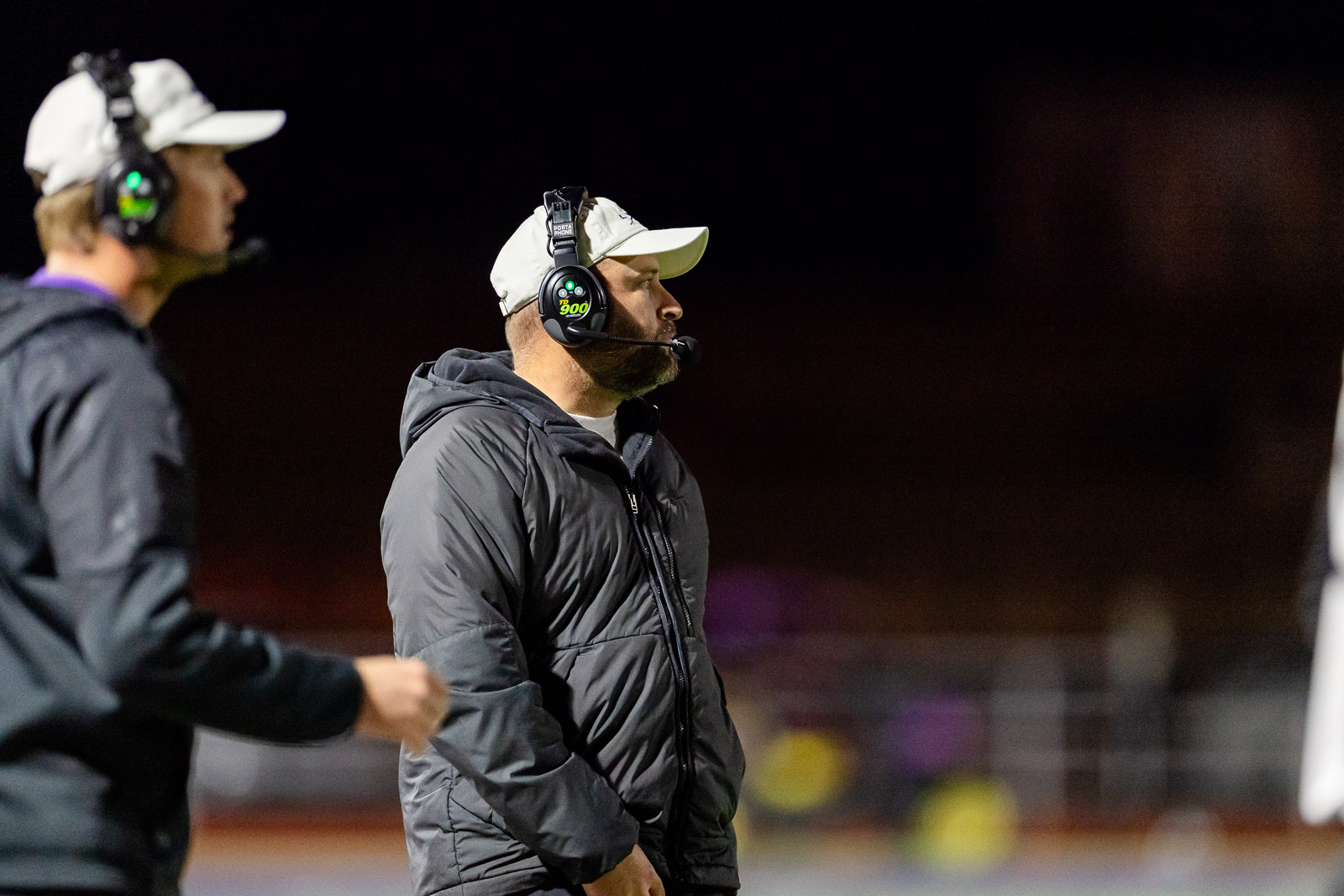 spearman-vs-panhandle-football-jpegJoe-pps-33 | Press Pass Sports Coach with white cap and gray jacket wearing a headset and microphone on a dimly lit track at night, facing forward.