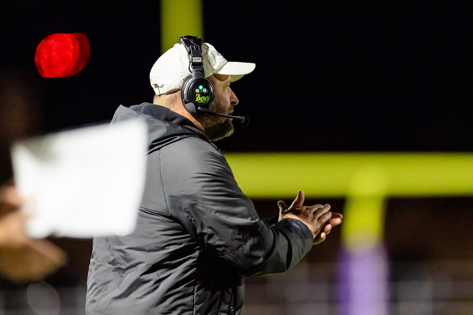 spearman-vs-panhandle-football-jpegJoe-pps-34 | Press Pass Sports Side view of a football coach in a white cap and gray jacket wearing a headset with mic, on a dimly lit sideline, clapping hands at night stadium.