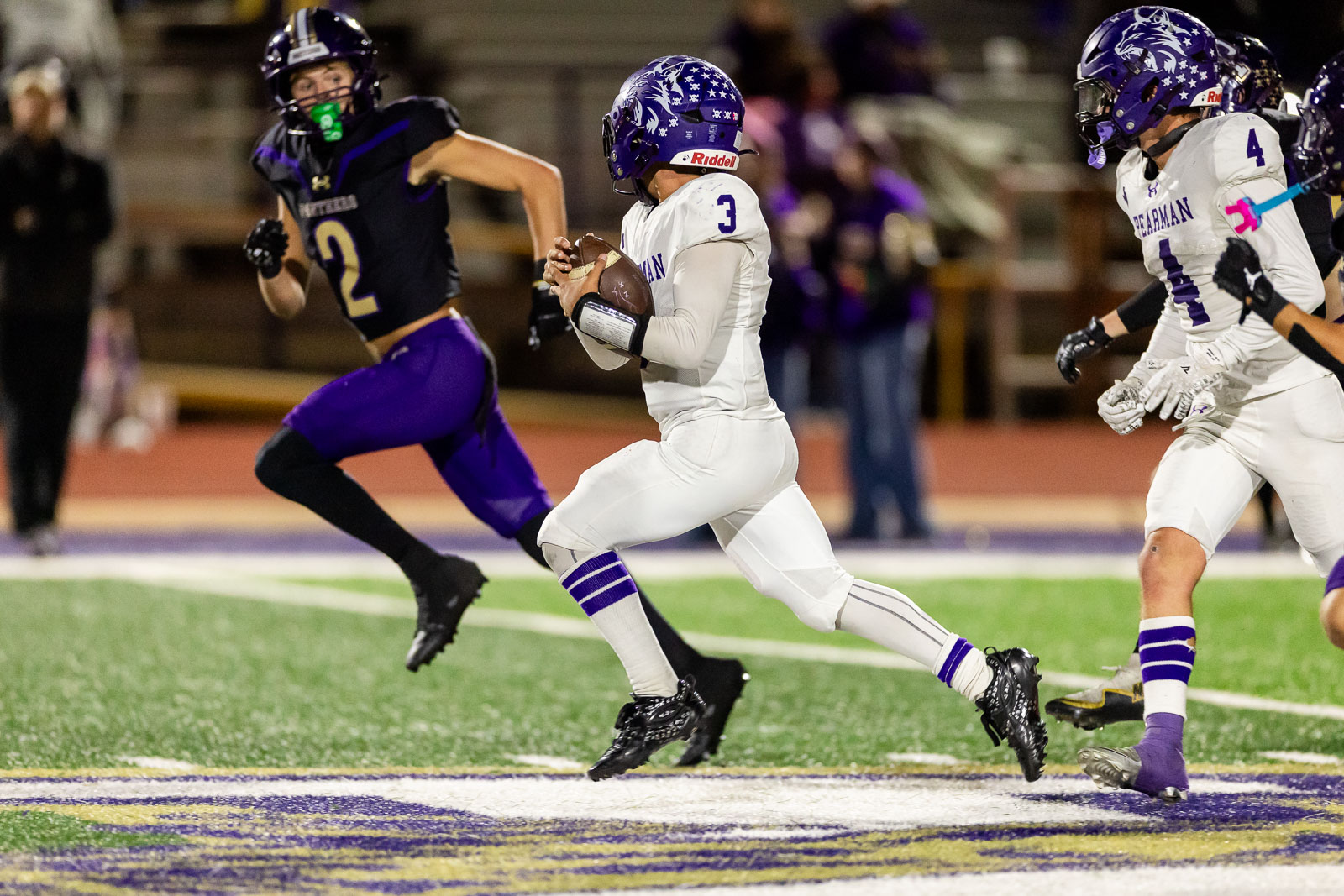 spearman-vs-panhandle-football-jpegJoe-pps-35 | Press Pass Sports White-uniform quarterback (number 3) runs with the football as a defender in purple (number 2) pursues him on a turf field under stadium lights.