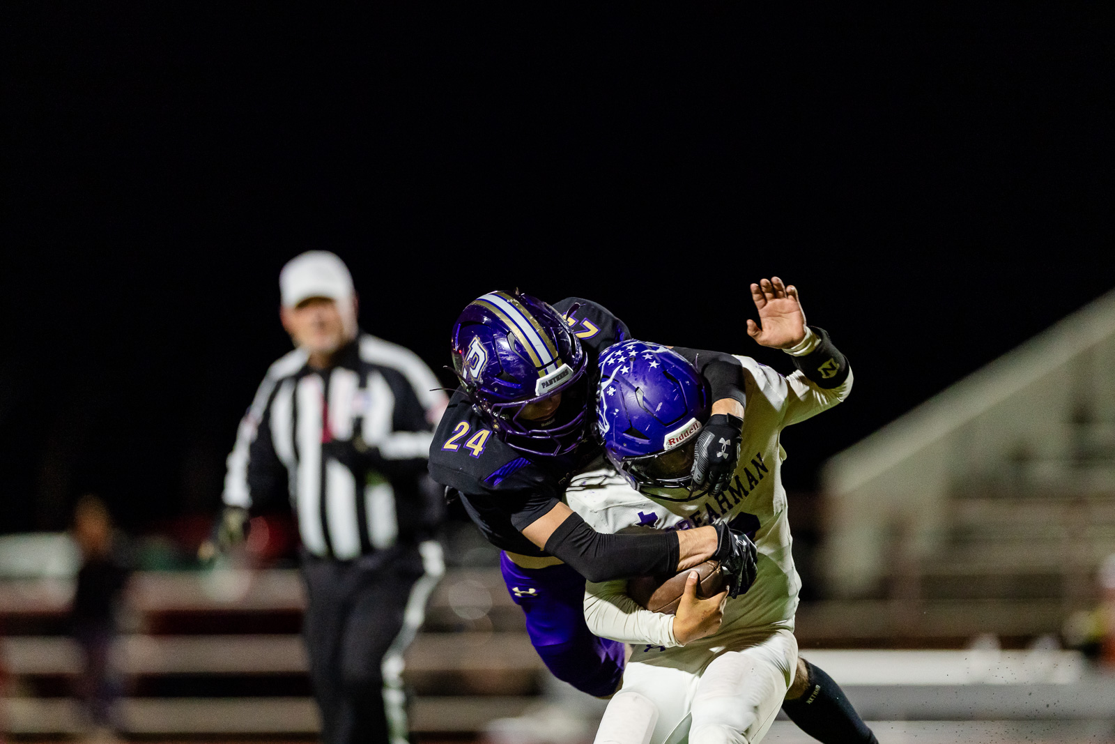 spearman-vs-panhandle-football-jpegJoe-pps-38 | Press Pass Sports Two football players clash in a nighttime tackle under stadium lights, one in purple and the other in white, with a referee in the background at left.