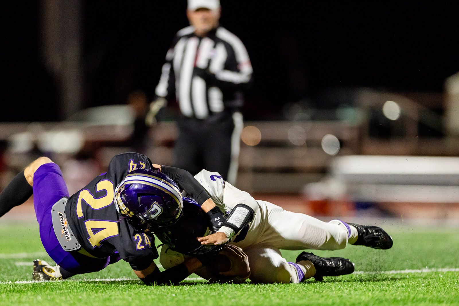 spearman-vs-panhandle-football-jpegJoe-pps-39 | Press Pass Sports Two football players collide on turf at night, one in purple and gold tackling a white-uniform opponent.