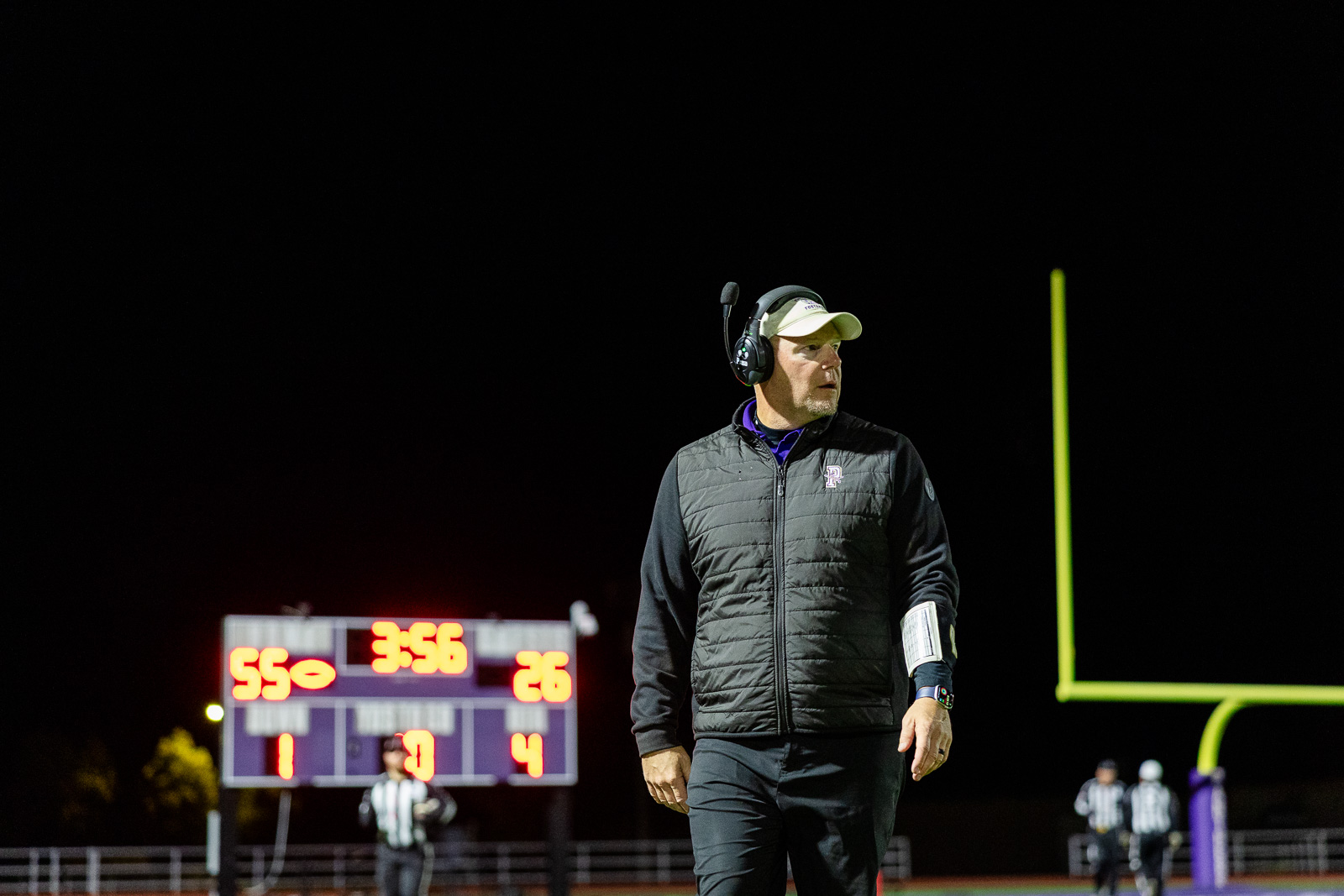 spearman-vs-panhandle-football-jpegJoe-pps-40 | Press Pass Sports Night football field: coach with headset and cap on the sideline, clipboard in hand, scoreboard glowing in the background