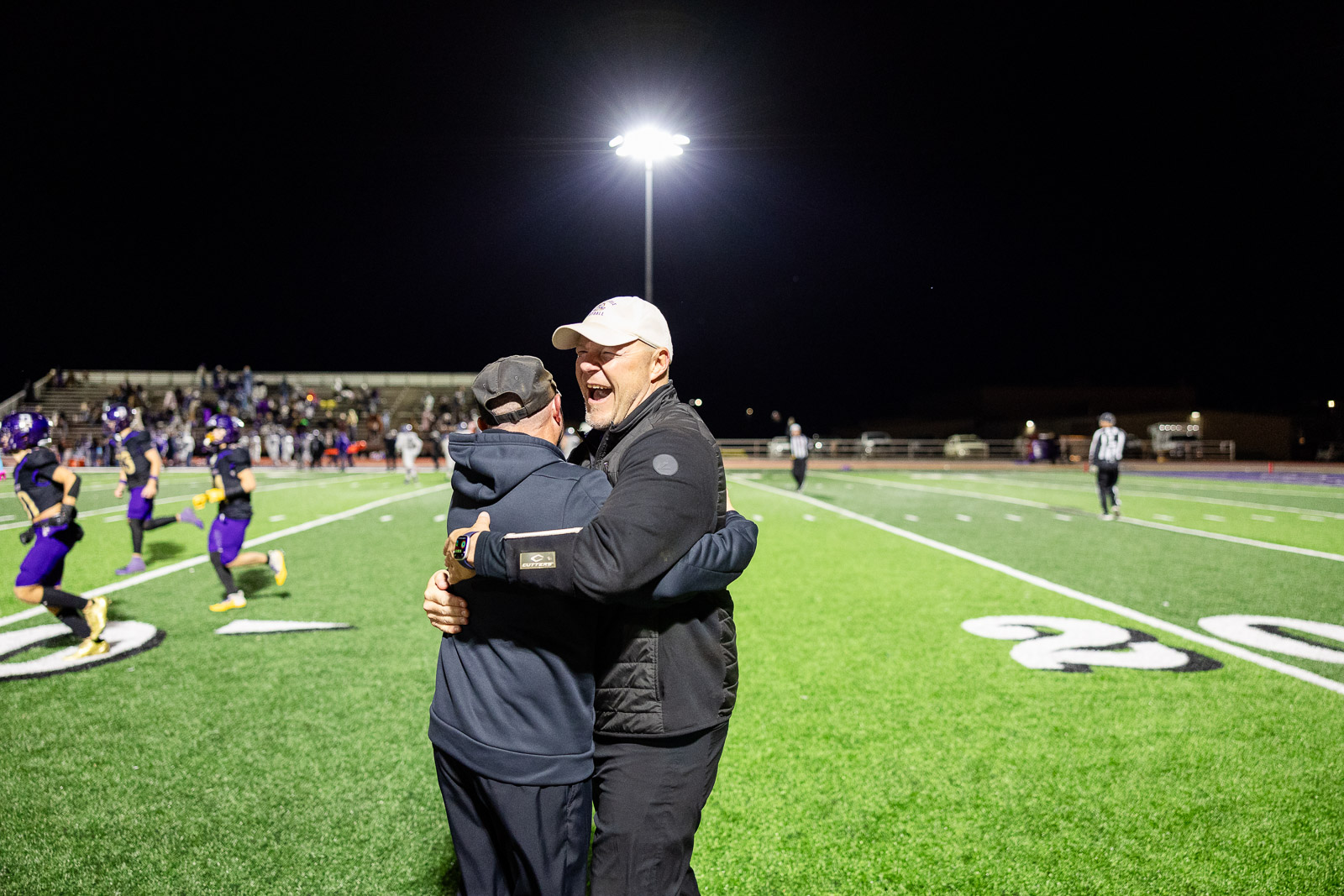 spearman-vs-panhandle-football-jpegJoe-pps-41 | Press Pass Sports Two coaches hug in the foreground on a lit football field at night, with players and bleachers in the background.
