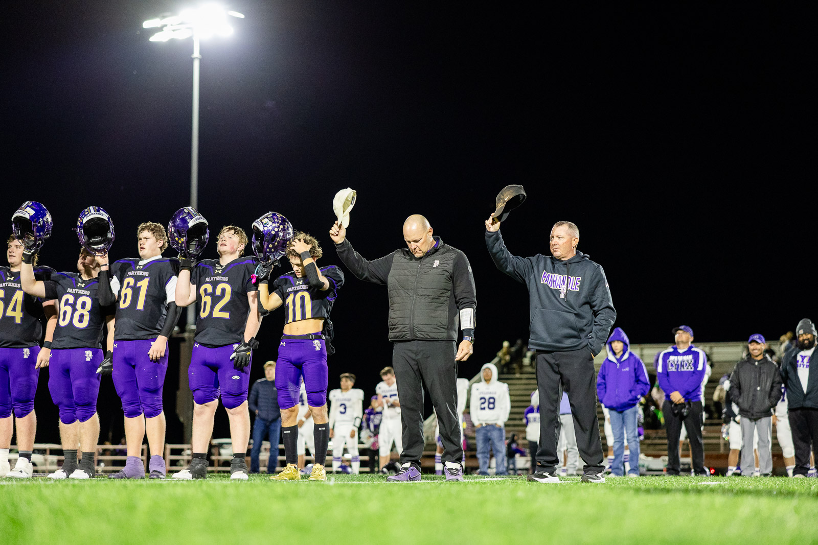 spearman-vs-panhandle-football-jpegJoe-pps-42 | Press Pass Sports High school football players in purple uniforms stand in a line on the field at night, lifting their helmets in salute beside two coaches holding hats under bright stadium lights.
