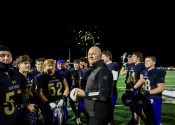 spearman-vs-panhandle-football-jpegJoe-pps-44 | Press Pass Sports Coach in black jacket stands with a high school football team on a bright field at night as fireworks glow behind them.