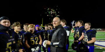 Coach in black jacket stands with a high school football team on a bright field at night as fireworks glow behind them.