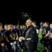 spearman-vs-panhandle-football-jpegJoe-pps-44 | Press Pass Sports Coach in black jacket stands with a high school football team on a bright field at night as fireworks glow behind them.