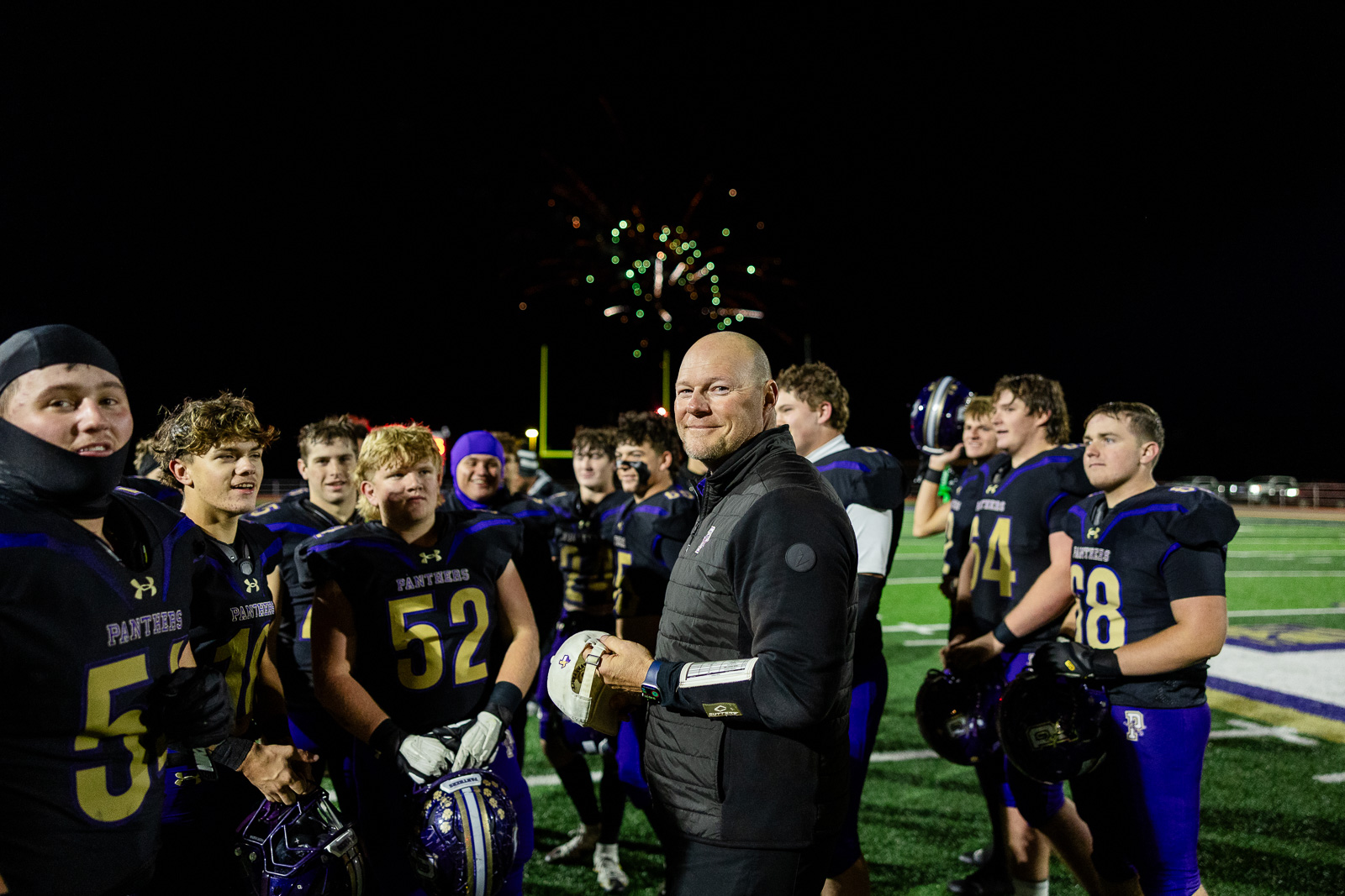 spearman-vs-panhandle-football-jpegJoe-pps-44 | Press Pass Sports Coach in black jacket stands with a high school football team on a bright field at night as fireworks glow behind them.