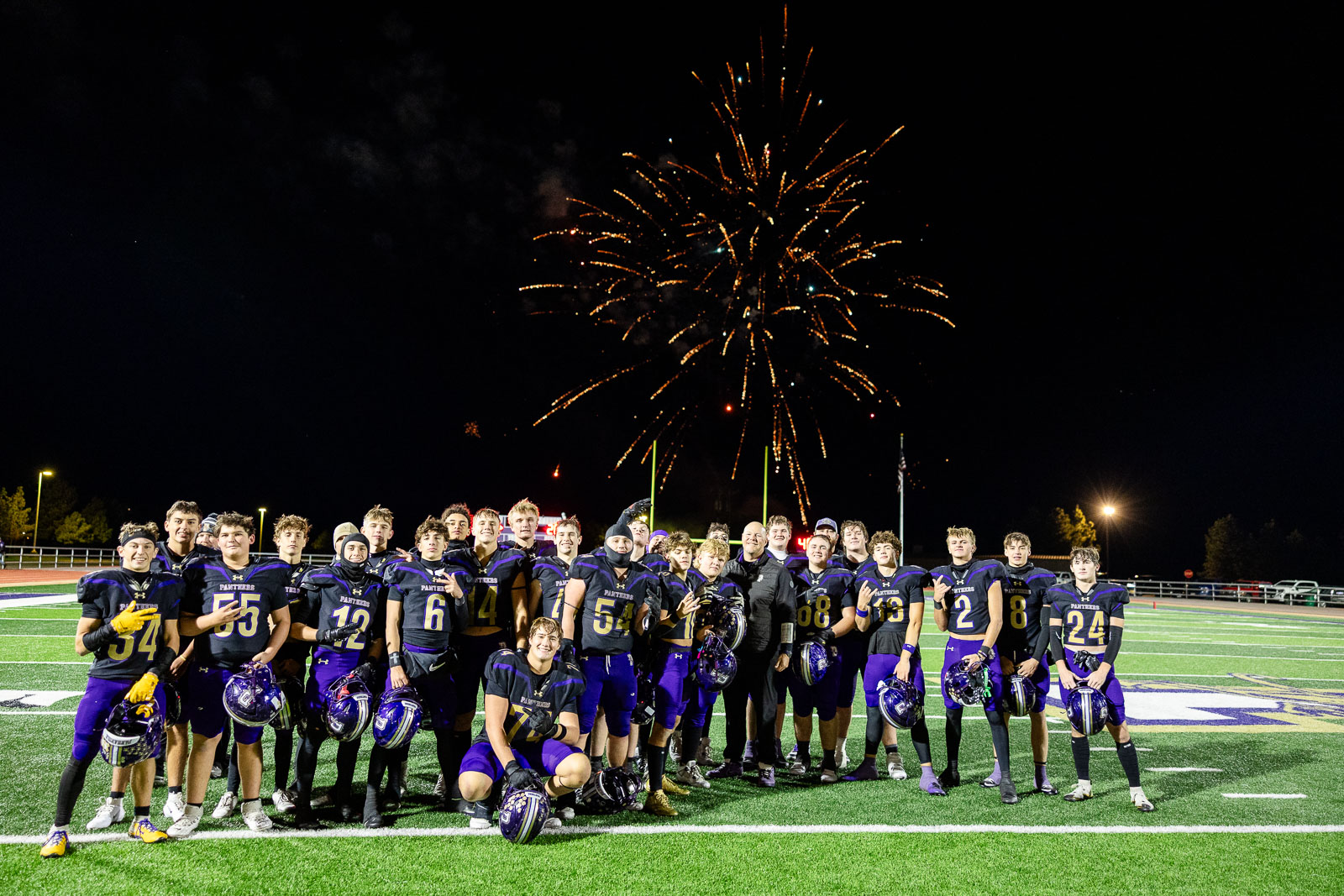 spearman-vs-panhandle-football-jpegJoe-pps-45 | Press Pass Sports Group of football players in purple and gold uniforms posing on a football field at night with fireworks exploding overhead; team members smile and hold helmets.