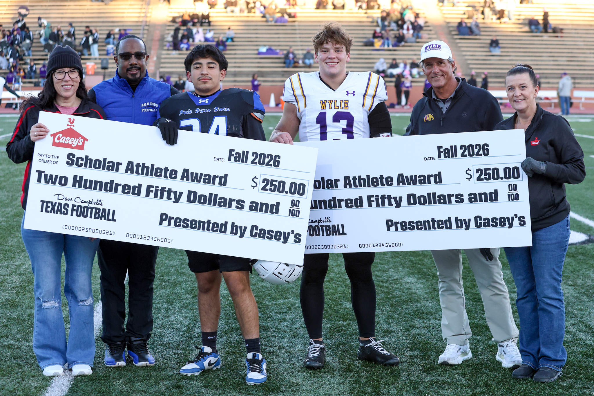 Six people on a football field pose with oversized checks reading'Scholar Athlete Award' for Fall 2026, $250 each.