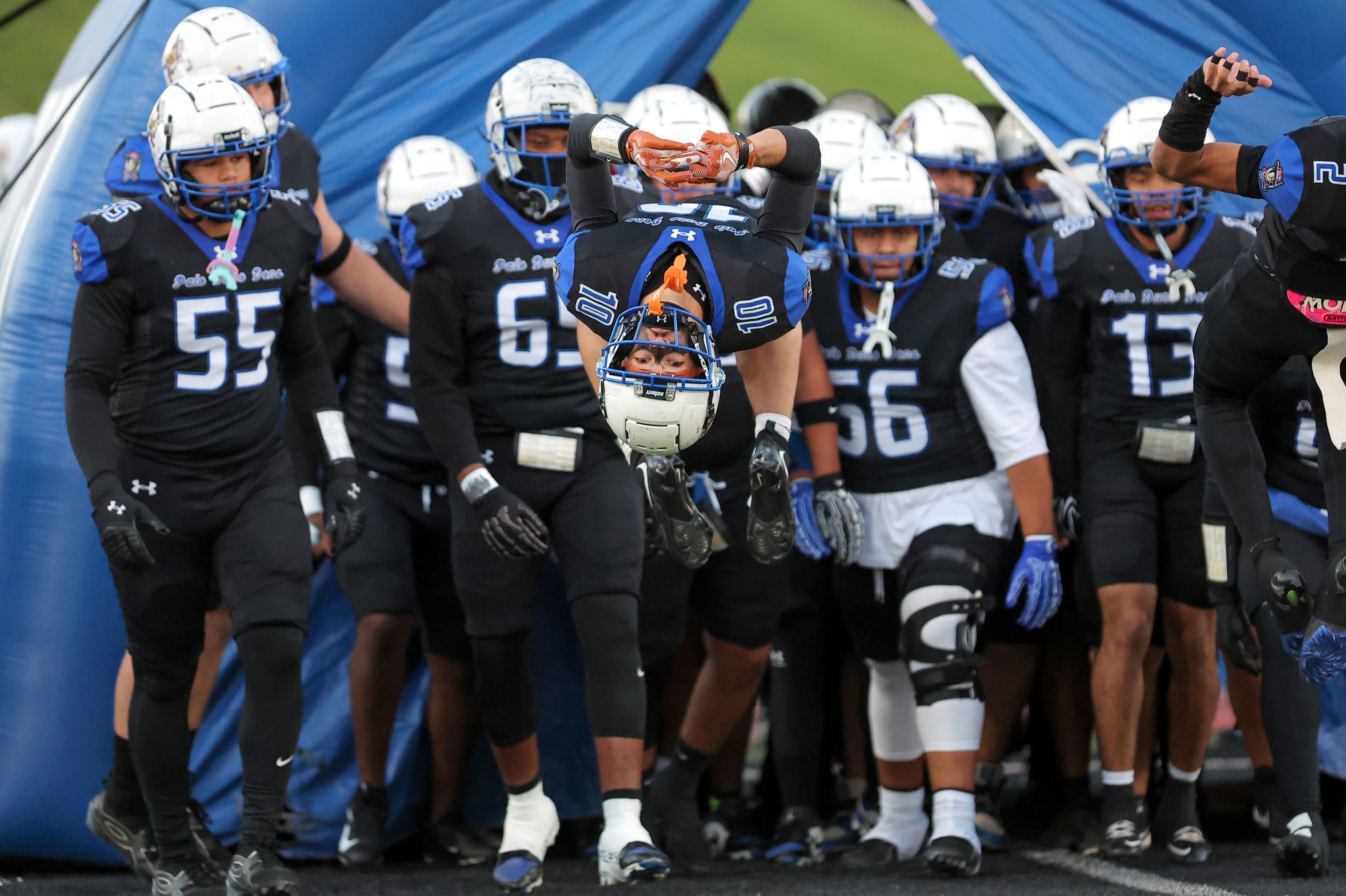 Football players in black-and-blue uniforms run through a blue tunnel toward the field; a central player performs a midair flip, arms folded overhead.