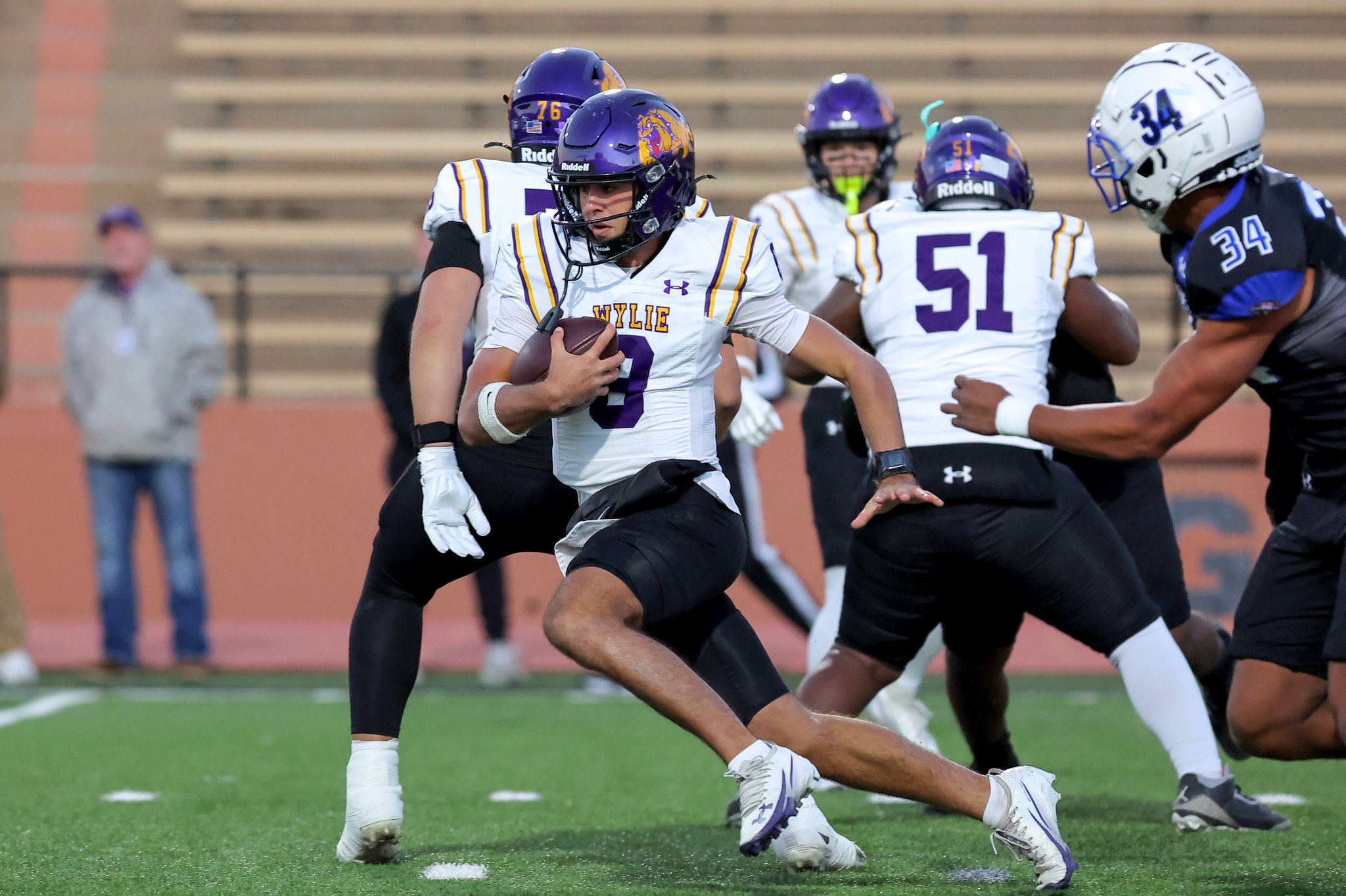 College football player in white and purple uniform carries the ball during a game, pursued by opponents in purple helmets and a blue-gray uniform.